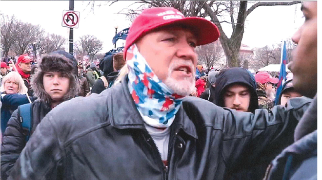 photo of a middle aged man wearing a red MAGA cap, multicolored gaiter, and black leather jacket, gesticulating in a crowd outside the US Capitol