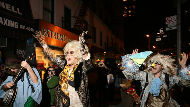 People in costumes dance down the street. A drag queen in a blonde wig and leopard-print gloves and jacket stands in the center.